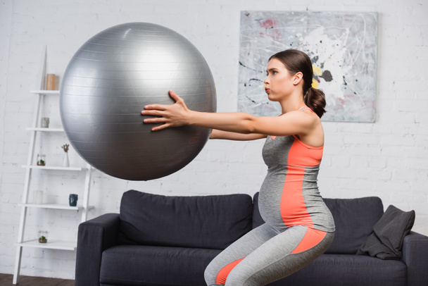 brunette femme enceinte respirant tout en faisant squat avec balle de fitness dans le salon  - Photo, image