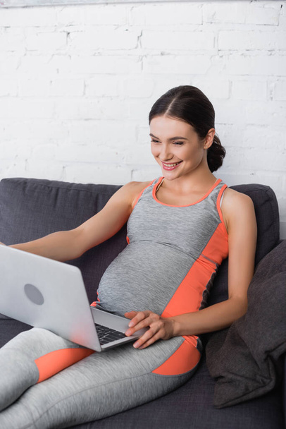 joyful, sportive and pregnant woman looking at laptop in living room - Fotoğraf, Görsel