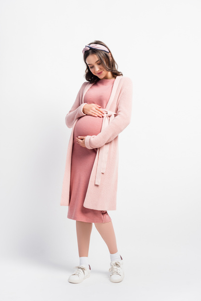 full length view of pregnant woman in cardigan and headband touching belly on white - Foto, Imagen