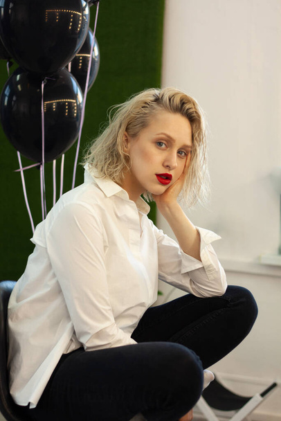 close-up portrait of beautiful young blonde woman with fashionable makeup and hair sitting bored in front of black balloons - Photo, Image
