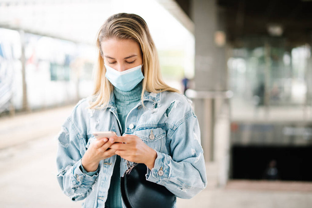 Young blonde woman wearing mask waiting in a bus station and using phone. - Valokuva, kuva