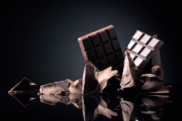 Μια σπασμένη σοκολάτα και κομμάτια μαύρης σοκολάτας σε μαύρο ανακλαστικό φόντο. Αντιγραφή χώρου. - Φωτογραφία, εικόνα