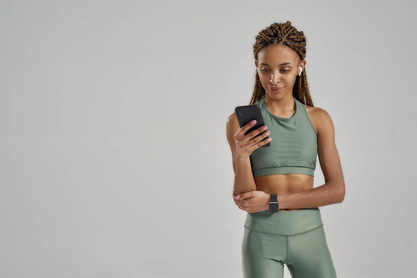 Молода змішана гоночна фітнес-леді в бездротових навушниках, використовуючи свій смартфон після тренування, стоячи в студії ізольовано на сірому фоні
 - Фото, зображення