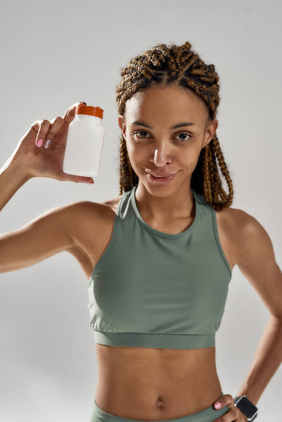 Vertical shot of a young mixed race woman in sportswear holding bottle with fitness nutrition supplements while standing isolated over grey background - Foto, imagen