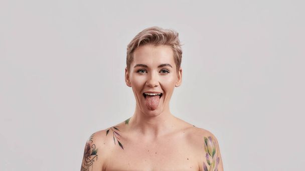 Portrait of a young attractive half naked tattooed woman with perfect skin showing her tongue to camera isolated over light background - Φωτογραφία, εικόνα