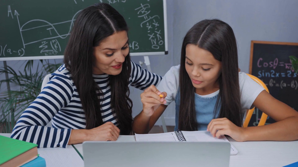 tyttö tekee läksyjä lähellä kannettavaa tietokonetta ja puhuu äidin kanssa - Materiaali, video