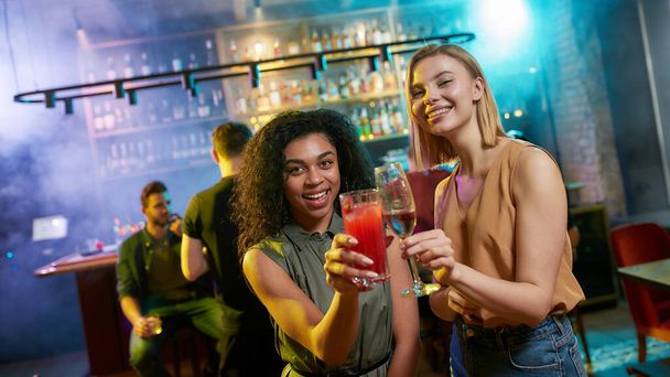 Attractive young women looking at camera while toasting, posing with cocktail in their hands. Friends celebrating, having fun in the bar - Photo, image