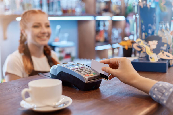 Customer paying through mobile phone over electronic reader in coffee shop, faceless customer holding card, smiling red haired waitress over counter. - Zdjęcie, obraz
