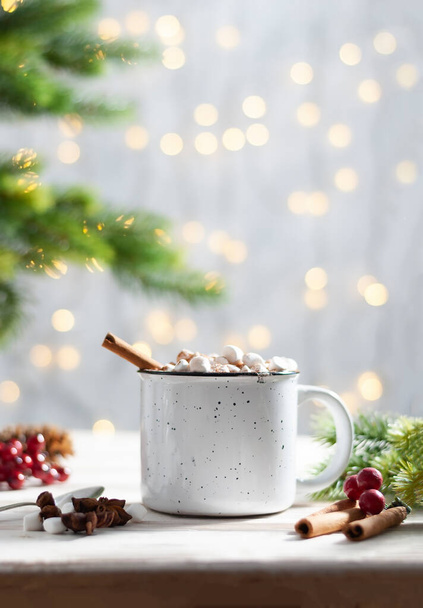 Christmas white cup with hot chocolate and marshmallows and red berries on with cinnamon sticks and fir xmas tree branches with beautiful garland lights. New year celebration vertical photo - Photo, Image