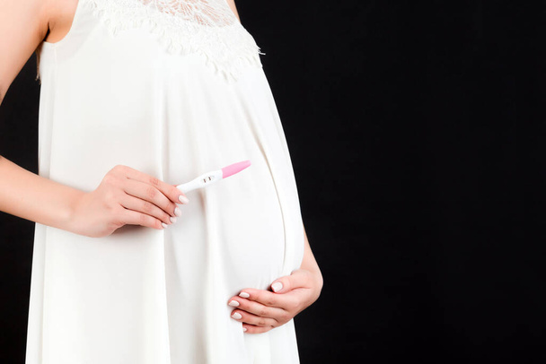 Großaufnahme einer schwangeren Frau im weißen Kleid mit positivem Schwangerschaftstest vor schwarzem Hintergrund. Mutterschaftskonzept. Kopierraum. - Foto, Bild