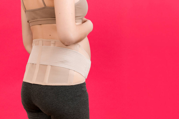 Vista posteriore della donna incinta che indossa la cintura di gravidanza su sfondo rosa con spazio per la copia. Primo piano del concetto di cintura di sostegno addominale ortopedica. - Foto, immagini