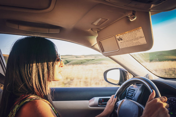 Young attractive girl in drivers seat looks straight and holds hands on steering wheel with nature outside the window in the background - Photo, Image