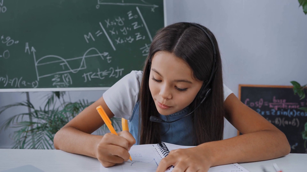 παιδί σε ακουστικά γράφοντας στο σημειωματάριο και χαιρετώντας το χέρι, ενώ online σπουδές  - Πλάνα, βίντεο