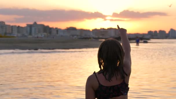 νεαρή κοπέλα δείχνει το ηλιοβασίλεμα στην παραλία - Πλάνα, βίντεο
