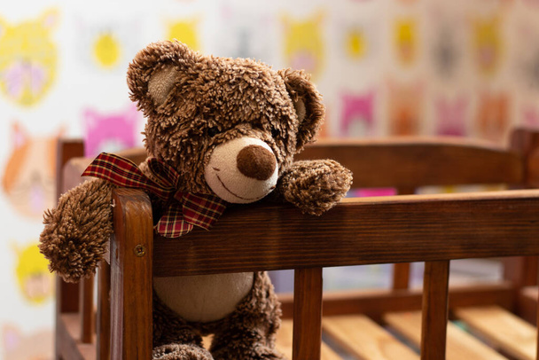 мягкий коричневый плюшевый мишка сидит в детской деревянной кровати в детской комнате на размытом фоне - Фото, изображение