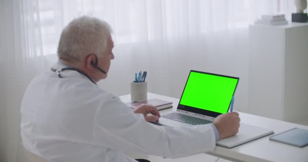 idős férfi orvos hallgat webinar vagy beteg laptopon zöld kijelző a chroma kulcs technológia, írás jegyzetek másolatot - Felvétel, videó