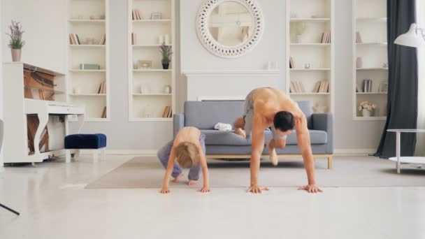 Active family doing morning exercise in living room for healthy lifestyle. Dad teaching cute small child boy how to do pushups. Fit sporty father and son play sports at home - 映像、動画