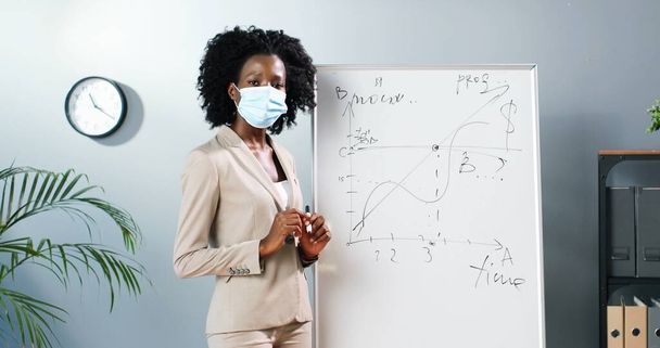 Young African American woman teacher in medical mask standing at board in classroom and telling physics or geometry laws to class. Pandemic concept. School during coronavirus. Educative lection. - Фото, зображення