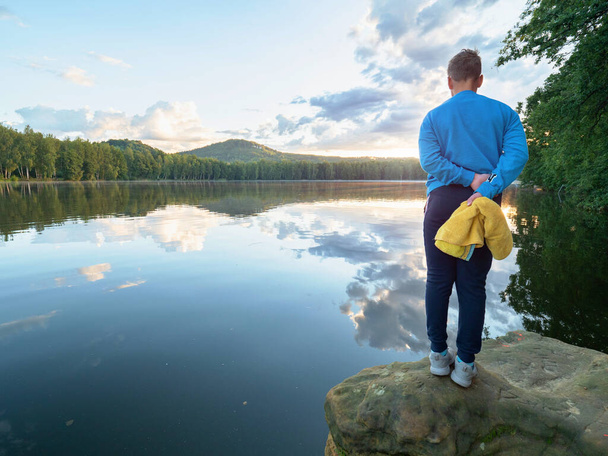 Синий мальчик в желтой рубашке и купальниках в руках стоит на скале над водой озера. Природный летний курорт. - Фото, изображение