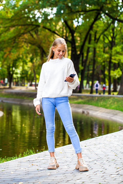 Full-length portrait of a young girl in a woolen sweater and blue jeans posing in an autumn park - Fotó, kép