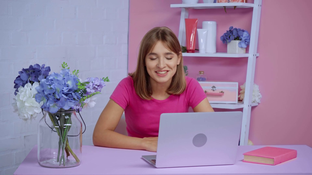 mujer alegre agitando la mano y hablando cerca de la computadora portátil y jarrón con flores en el escritorio - Metraje, vídeo