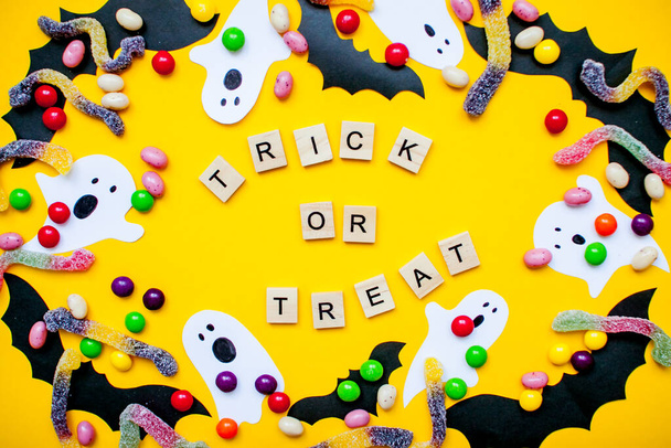 inscription from wooden blocks trick or treat and frame made of paper homemade bats and paper ghosts and multicolored candies and worms from gummy on a bright yellow background - Photo, image