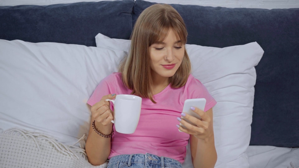 mujer joven sosteniendo la taza, bebiendo café y usando el teléfono inteligente en la cama - Metraje, vídeo