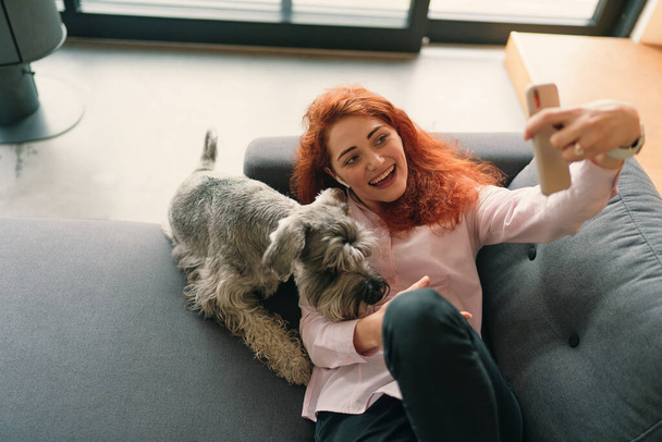 Girl with red hair and charming smile lying on sofa and making selfie with her pet - Foto, imagen
