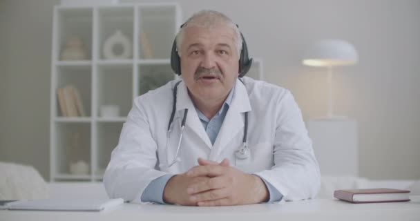 cardiólogo masculino está consultando en línea, sentado en la oficina de la clínica y respondiendo al paciente por videochat en el ordenador portátil - Imágenes, Vídeo