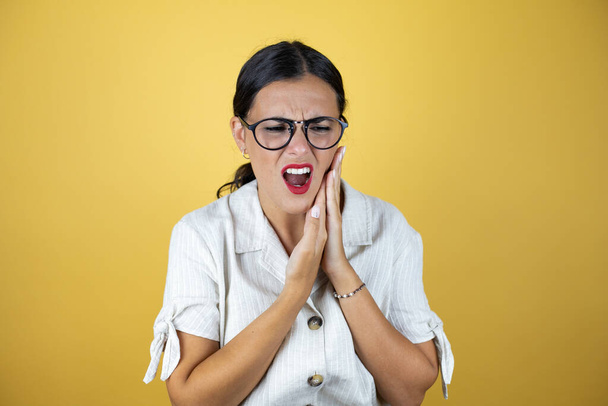 Mooie vrouw over gele achtergrond aanraken mond met de hand met pijnlijke uitdrukking als gevolg van kiespijn of tandheelkundige ziekte op tanden. - Foto, afbeelding