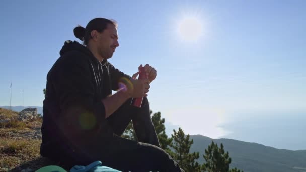 Nuori turisti nauttii merinäköalasta korkealta vuorelta. Mies juo teetä termospullosta istuessaan kalliolla vuorilla nousevan auringon säteillä. - Materiaali, video