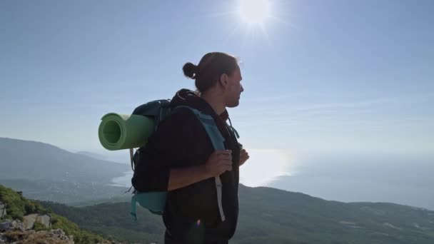 Ένας νεαρός, αρσενικός τουρίστας σκαρφαλώνει στα βουνά. Ένας ταξιδιώτης με σακίδιο στην πλάτη στέκεται στην κορυφή του βουνού. Θέα στη θάλασσα στις ακτίνες του ανατέλλοντος ήλιου. - Πλάνα, βίντεο