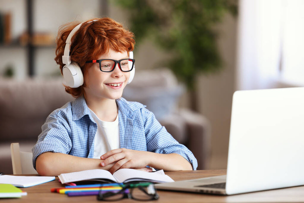 Happy Cute рыжий мальчик в наушниках улыбается во время общения с учителем через видео чат приложение во время онлайн-образования в доме - Фото, изображение