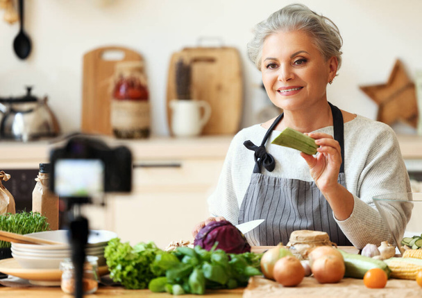 正成熟した女性でエプロン保持新鮮なスカッシュながらビデオレシピの健康的な菜食主義者の料理で居心地の良い家庭用キッチン - 写真・画像