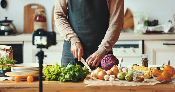 Nierozpoznawalny mężczyzna w fartuchu kroi warzywa podczas nagrywania wideo do gotowania vlog w kuchni w hom - Zdjęcie, obraz