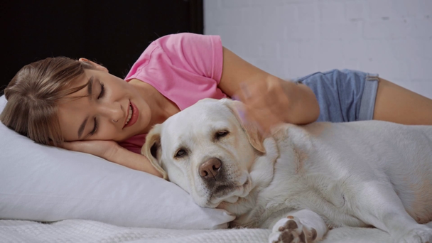 zadowolona kobieta leżąca na poduszce i przytulająca golden retriever - Materiał filmowy, wideo