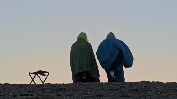 Een groep jonge vrienden ontmoet de zonsopgang op de berg. Slaperige toeristen in slaapzakken, zittend op stoelen op een klif. Een prachtig uitzicht op de stad aan de voet van de berg in de stralen van de rijzende zon. - Video