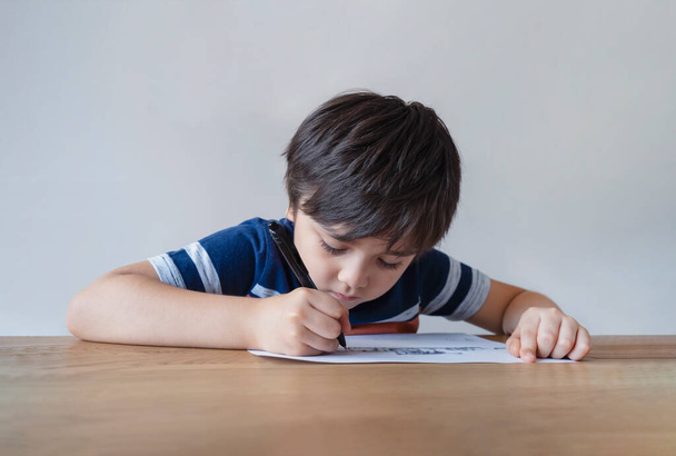 Portrait d'un écolier assis seul faisant ses devoirs, un enfant tenant un stylo noir dessinant et écrivant sur du papier blanc sur une table, concept d'école primaire et d'enseignement à domicile - Photo, image