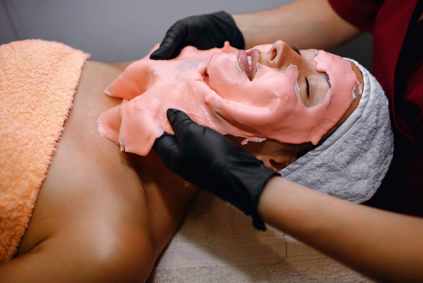 Una chica morena toma procedimientos en un salón de spa, una máscara rosa aplicada en una capa gruesa es removida por un maestro en guantes negros, limpiando la piel de la suciedad, monitoreando la pureza de la cara - Foto, imagen