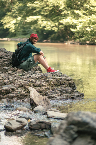 Tourist guy with backpack sitting near lake in a wild summer wood. Expedition - Foto, imagen