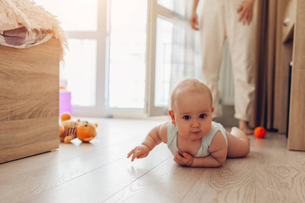 Infant crawling on floor in bedroom while mom watches. Little baby having fun at home. Kid looks at camera - Photo, Image