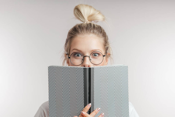 Blonde woman with shocked eyes hiding behind an open book and looking at camera. - Photo, Image