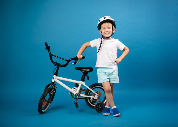 Un niño pequeño con la bicicleta de un niño y el casco protector está de pie sobre un fondo azul y mira a la cámara. Foto de estudio para artículos sobre seguridad y deportes infantiles y actividad. - Foto, imagen