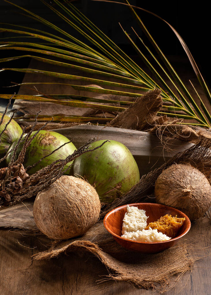 Cocos verdes abiertos y cocos maduros con cocada dulce (dulce brasileño) sobre fondo de madera y hojas de coco secas - Foto, Imagen