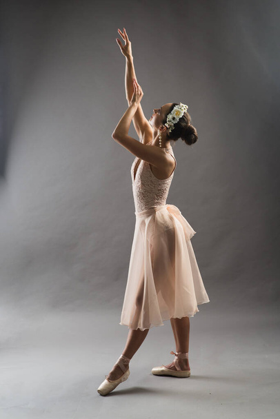 молодая красивая балерина в бежевом купальнике позирует на пуантах на светло-сером фоне студии - Фото, изображение