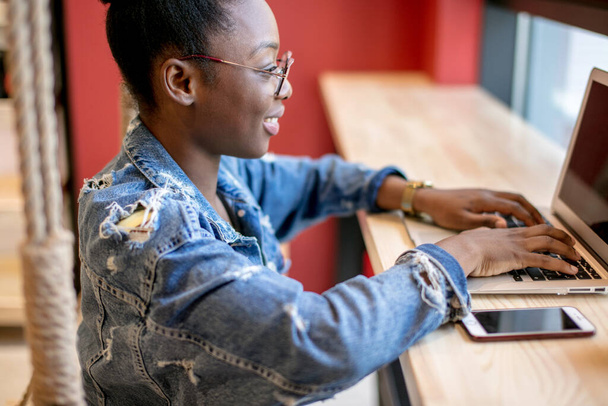 African woman in denim jacket sitting near the window and using laptop in cafe - Foto, immagini