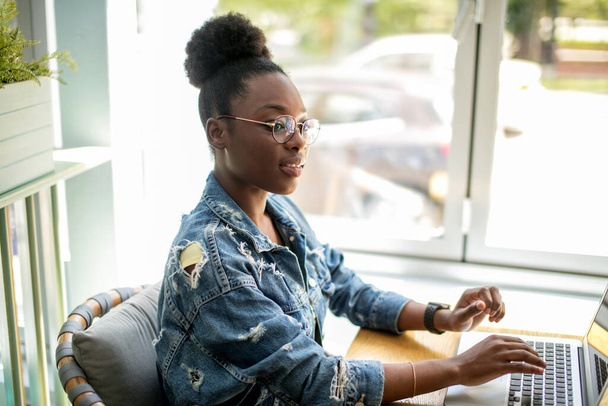 African woman in denim jacket sitting near the window and using laptop in cafe - Foto, imagen