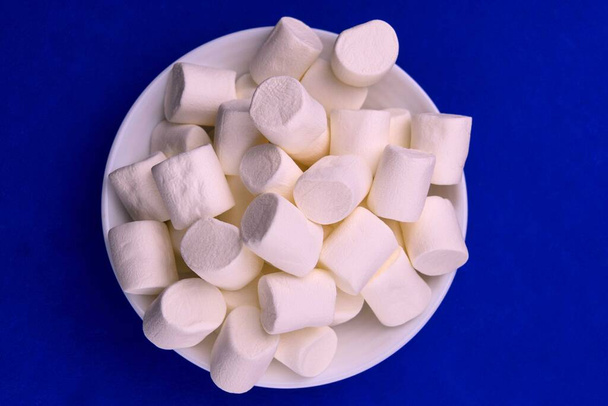 Bílý načechraný bílý marshmallow v bílém talíři na tmavomodrém pozadí. Velký horní pohled na makro marshmallow - Fotografie, Obrázek
