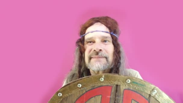 Pink background. Mature man, crazy viking with long hair and beard rejoices and waves his hand hiding behind shield. Joke humor - Footage, Video