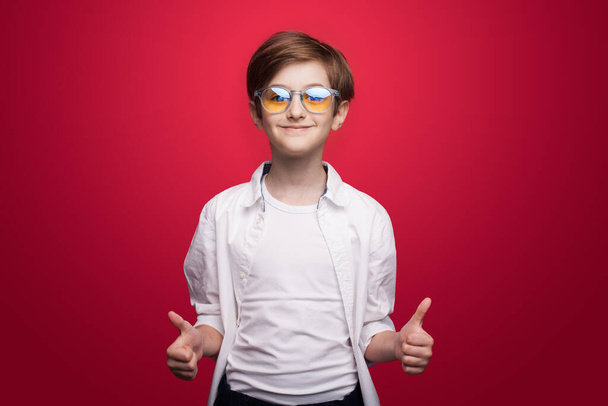Tyytyväinen valkoihoinen poika, jolla on lasit ja valkoinen paita elehtivät kuin merkki ja hymy kameralle punaisella studion seinällä - Valokuva, kuva
